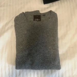 Grå stickad tröja från Oscar Jacobson - Mysig grå stickad tröja från Oscar Jacobson i klassisk modell. Tröjan har rund halsringning, ribbade muddar och är långärmad. Perfekt för lager-på-lager och passar till både jeans och chinos.