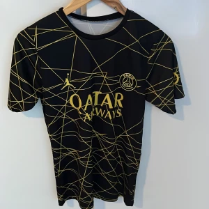 PSG tröja - Snygg PSG fotbollströja med Mbappé 7 på ryggen. Svart bas med gula linjer i ett unikt mönster, Qatar Airways-logga och Jordan-märke på bröstet. Tillverkad i lätt och ventilerande material, perfekt för träning eller match.