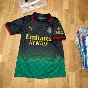 AC Milan x Off-White x Puma fotbollströja - AC Milan 2025 special edition fotbollströja från Off-White och Puma. Svart och grön med gula tryck, röda detaljer på krage och ärmslut. Kortärmad, med tryck 'JOÃO FÉLIX 79' på ryggen. Tillverkad i lätt funktionsmaterial med diskret mönster.