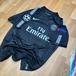 PSG Neymar Jr svart matchtröja Nike L - Svart PSG fotbollströja från Nike med Neymar Jr och nummer 10 på ryggen. Tröjan har korta ärmar, Champions League och Respect-patchar, samt Fly Emirates-tryck på bröstet. Tillverkad i Dri-FIT-material som andas, med diskreta grafiska mönster och vita detaljer.