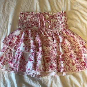 Vit och rosa blommig volangkjol - Supersöt kort kjol med volanger och smock i midjan. Vit botten med rosa blommor och knytband framtill. Perfekt för sommarens alla äventyr och ger en riktigt fräsch vibe. Funkar även att ha som en söt topp 💘💘