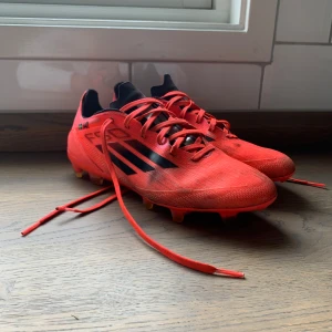 Röda Adidas fotbollsskor PRO - Snygga röda Adidas fotbollsskor med svarta ränder och svarta detaljer. Skorna har snörning och är designade för att ge bra grepp på planen. Lätta och smidiga med syntetmaterial och klassisk Adidas-look. Perfekta för dig som vill sticka ut på fotbollsplanen.