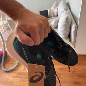 Löparskor (ASICS) - nya och oanvända. Nypris 1800 kr. - Snygga svarta Asics sneakers med turkosa och vita detaljer. Ovandelen är i ventilerande mesh och sulan är vit och tjock för extra komfort. Skorna har snörning och Ortholite-innersula för skön passform. Perfekta för dig som gillar sportig stil.