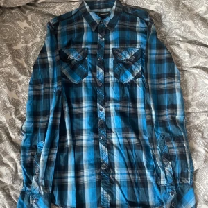 Blå rutig skjorta med knappar - Snygg blå och svart rutig skjorta med långa ärmar och knappar framtill. Skjortan har två bröstfickor med lock och knapp, klassisk krage och en avslappnad passform. Perfekt för dig som gillar en cool och avslappnad stil. Säljer många skjortor så köp gärna flera i en bundle!