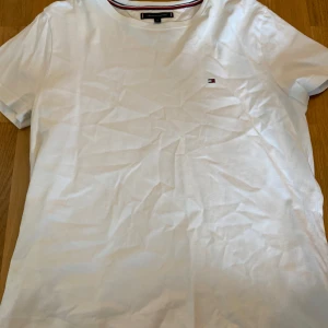 Vit t-shirt från Tommy Hilfiger i strlM - Klassisk vit t-shirt från Tommy Hilfiger. T-shirten har korta ärmar, rund halsringning med snygg randig detalj och liten broderad logga på bröstet. Kan mötas upp i Trollhättan eller Alingsås. Kan skickas men då tillkommer en fraktkostnad.