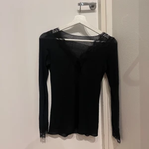 Svart långärmad topp med spets - Supersnygg svart långärmad topp med v-ringning och spetsdetaljer vid halsringning och ärmslut. Figurnära passform som framhäver siluetten. Perfekt att styla med jeans eller kjol för en trendig look.