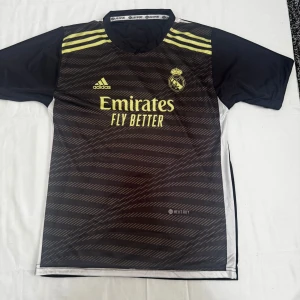 Real Madrid Vinicius Jr. matchtröja Adidas - Snygg svart Real Madrid fotbollströja från Adidas med gula detaljer och tryck. Tröjan har korta ärmar, randigt mönster på framsidan, klubbmärke och sponsortryck. På ryggen står det VINIJR. och nummer 20. Tillverkad i lätt och ventilerande material.