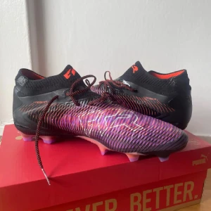 Puma Future Z fotbollsskor svart/lila - Säljer ett par Puma Future Z fotbollsskor i svart och lila med coola orange detaljer. Skorna har snörning, stickad strumpliknande krage och mönstrad ovandel för extra grepp. Sulan har lila och rosa dobbar för bästa fäste på planen. Perfekta för dig som vill sticka ut på matchen!