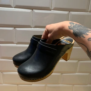 Svarta träskor med klack från swedish hasbeens - Klassiska svarta träskor från swedish hasbeens med rund tå och snygg träklack. Ovandelen är i svart skinn och sulan i ljust trä, vilket ger en stilren och tidlös vibe