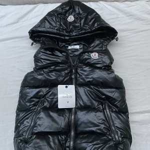 Svart dunväst från Moncler med huva - Säljer en svart, glansig dunväst från Moncler med huva och dragkedja framtill. Västen har två snedställda fickor med dragkedja och klassisk Moncler-logga på bröstet. Perfekt för lager-på-lager och riktigt snygg till streetwear.
