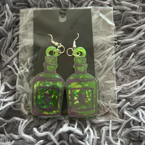 Gröna örhängen i form av giftflaskor - Unika örhängen formade som gröna giftflaskor med lila dödskallemotiv. De är genomskinliga med glittriga inslag i grönt och gult, och har silvriga krokar. Perfekta för dig som vill sticka ut med en cool och lekfull stil.