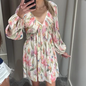 Blommig kortklänning med v-ringning - Superfin kortklänning med blommigt mönster i rosa, grönt och vitt. Klänningen har v-ringning, långa puffärmar med snörning vid ärmslut och ett luftigt, plisserat tyg. Perfekt för sommarens alla tillfällen!