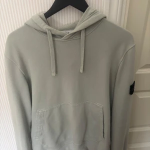 Mint grön hoodie från Stone Island - Säljer en stilren Mintgrön hoodie från Stone Island med klassisk patch på vänster ärm. Tröjan har huva med snörning och en stor magficka framtill. Perfekt för dig som gillar streetwear och vill ha något bekvämt och snyggt i garderoben.