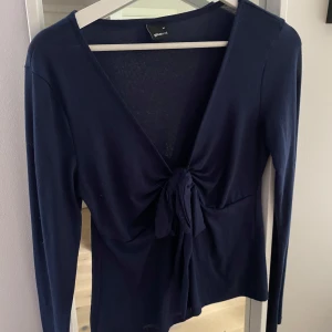 Mörkblå omlottopp från Gina Tricot - Snygg mörkblå omlottopp från Gina Tricot med lång ärm och djup v-ringning. Toppen har en knytning framtill som ger en cool och trendig look. Perfekt att styla med jeans eller kjol för en enkel men chic outfit.