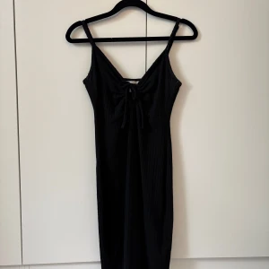 Svart ribbad klänning H&M XS - Svart ribbad kortklänning från H&M i storlek XS. Klänningen har smala axelband, v-ringning och en söt knytning framtill. Den är figurnära och har en enkel, stilren look som passar perfekt till många olika tillfällen.