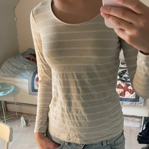 Beige randig långärmad tröja BASIC - Säljer en beige långärmad tröja från BASIC med tunna horisontella ränder i en ljusare nyans. Tröjan har rund halsringning och är i ett mjukt bomullsmaterial som sitter tajt mot kroppen. Perfekt för lager-på-lager eller som den är till jeans.