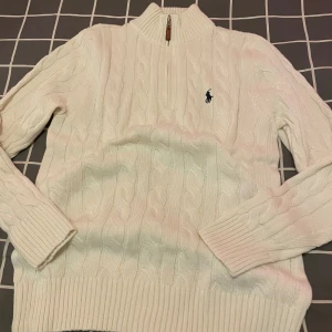 Vit cardigan stickad tröja Polo Ralph Lauren - Vit kabelstickad tröja från Polo Ralph Lauren med halv dragkedja och hög krage. Klassisk broderad logga på bröstet och ribbade muddar. Perfekt för lager-på-lager och snygg till jeans eller kjol.