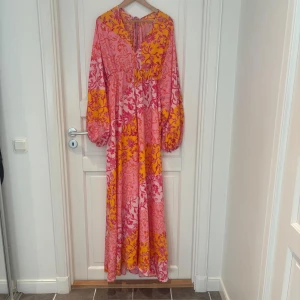 Färgstark långklänning med blommönster. 44/46 - Långklänning i bohemisk stil med rosa och orange botten, täckt av röda och rosa blommor. Klänningen har långa, vida ärmar, v-ringning och snörning i ryggen. Luftigt material, perfekt för varma dagar. Snygg och unik statement-piece för dig som gillar färg.