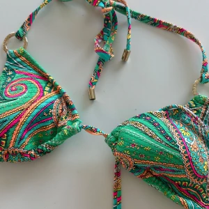 Färgglad paisley bikini med snörning - Superfin bikini med paisleyinspirerat mönster i grönt, gult, orange och rosa. Triangelformad överdel med knyt i nacken och ryggen, samt guldfärgade ringdetaljer. Perfekt för stranden eller poolen när du vill sticka ut lite extra.