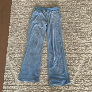 Blå velourbyxor från Juicy Couture - Mysiga ljusblå velourbyxor från Juicy Couture med broderad JC-logga på fickan, snörning i midjan och bakfickor med knapp. Byxorna har en loose passform och raka ben, perfekta för en chill och trendig look.
