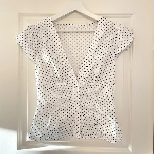 Vit prickig blus med v-ringning - Supersöt vit blus med svarta prickar och djup v-ringning. Korta ärmar och knappar framtill ger en feminin och trendig look. Perfekt till jeans eller kjol för en fräsch stil. Lätt och luftig, passar perfekt till varmare dagar. Köptes här på plick men var tyvärr lite stor så därför jag säljer💓pris kan diskuteras vid snabbt köp!