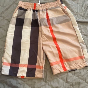 Burberry shorts - Snygga shorts med rutigt mönster i beige, svart, vitt och rött. De har elastisk midja med snörning, glansigt material som känns svalt. Perfekta för sommaren och har fickor i sidorna. Passformen är normal och längden går till knäna. Priset kan diskuteras .Storleken är S men funkar för M oxå