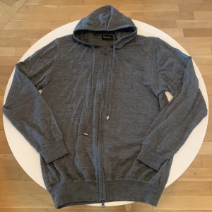 Grå hoodie från Sand Copenhagen - Mörkgrå hoodie från Sand Copenhagen med dragkedja framtill och snörning i huvan. Tröjan är tillverkad i ett mjukt, stickat material och har långa ärmar samt ribbade muddar. Perfekt för en chill och stilren look.