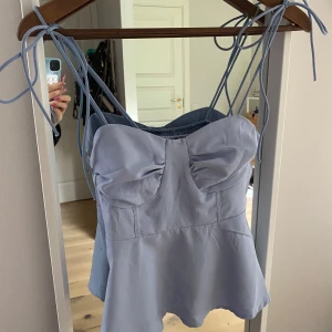 Ljusblå linnetopp med knytband - Supersöt ljusblå linnetopp med smala axelband som knyts upptill. Toppen har en söt rynkad detalj vid bysten och smockad rygg för extra komfort. Lätt utsvängd nertill och perfekt för varma dagar.