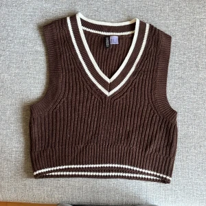 Brun stickad väst från H&M Divided XS - Säljer en brun stickad väst från H&M Divided i storlek XS. Västen har v-ringning och vita ränder vid halsringning och nederkant. Den är ribbstickad och har en klassisk, croppad passform som är perfekt att styla över en skjorta eller t-shirt.