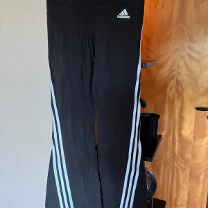Svarta Adidas byxor med vita ränder - Svarta Adidas byxor med klassiska vita ränder längs benen och Adidas-logga framtill. Byxorna har bootcut-modell med utsvängda ben och är tillverkade i mjukt material som passar perfekt till träning eller chill. Elastisk midja för skön passform. Ca 60 cm midjemått och 91 cm topp till tå.
