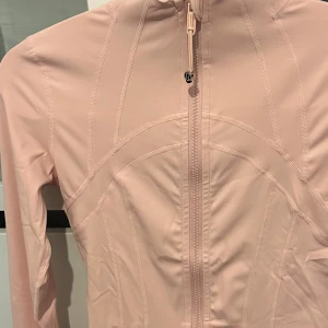 Ljusrosa träningsjacka från Lululemon - (PRIS KAN DISKUTERAS VID SNABBAFFÄR) - Supersnygg ljusrosa träningsjacka från Lululemon med figurnära passform, hel dragkedja framtill och höga fickor med dragkedja. Jackan har långa ärmar och är tillverkad i ett mjukt, stretchigt syntetmaterial som andas. Perfekt för gymmet eller powerwalks.