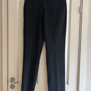 Svarta kostymbyxor med rak passform - Snygga svarta kostymbyxor med klassisk rak passform och pressveck. Byxorna har diskreta sidfickor och en stilren midja utan synliga detaljer. Perfekta för en clean och dressad look. Materialet känns mjukt och följsamt.