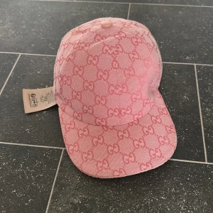 Rosa Gucci keps med GG-mönster - Snygg rosa keps från Gucci med ikoniskt GG-mönster över hela kepsen. Justerbar rem baktill i matchande rosa och böjd skärm. Tillverkad i bomullstyg med exklusiv känsla och tydlig logga på insidan. Perfekt för dig som vill sticka ut med en lyxig accessoar.