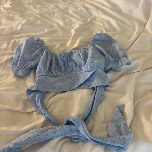 Blå omlott blus med puffärmar - Supersöt ljusblå blus med broderade hål och puffiga korta ärmar. Toppen har omlottknytning i midjan och volangdetaljer vid ärmslut. Perfekt för sommarens alla tillfällen och ger en romantisk vibe.