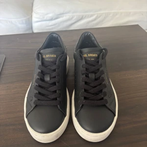 Svarta Axel Arigato sneakers i skinn - Stilrena svarta sneakers från Axel Arigato med vit sula och diskret guldfärgad logga på plösen. Skorna har rund tå, svarta skosnören och är tillverkade i slätt skinn. De är endast använda en gång så fortfarande i nyskick. Box saknas men skickas med dustbag! Storlek 36, pris går att diskutera