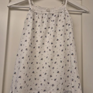 Vit blommig linnetopp från Newbie - Supersöt vit linnetopp från Newbie med små blå och beige blommor. Toppen har smala axelband, spetsdetaljer och en liten rosett framtill. Perfekt för varma dagar och ger en romantisk vibe med sitt luftiga bomullsmaterial.