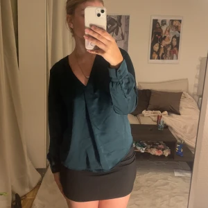 Mörkgrön omlottblus med v-ringning - Snygg mörkgrön omlottblus med v-ringning och långa ärmar. Blusen har en lätt glansig finish och knappar vid ärmsluten, vilket ger en elegant touch. Perfekt att styla med kjol eller jeans för en trendig look.