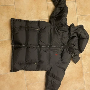 Svart pufferjacka från Polo Ralph Lauren - Svart pufferjacka från Polo Ralph Lauren med röd broderad logga på bröstet. Jackan har flera dragkedjeförsedda fickor, huva med snörning och en klassisk quiltad look. Perfekt för kalla dagar när du vill hålla stilen och värmen.