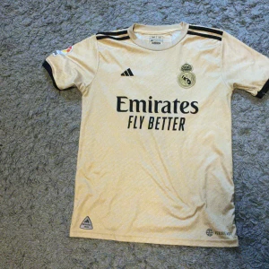 Real Madrid Vini Jr. matchtröja Adidas - Snygg Real Madrid fotbollströja i ljusbeige med svarta detaljer och tryck. Adidas-logga, klubbmärke och sponsor framtill, samt VINI JR. 20 på ryggen. Korta ärmar, klassisk passform och tillverkad i lätt funktionsmaterial. Pris kan diskuteras vid snabb affär 