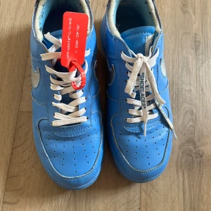 Nike Air Force 1 x Off-White blå - Säljer ett par Nike Air Force 1 x Off-White i blått skinn med silvriga swoosh-detaljer och vit snörning. Skorna har den ikoniska röda zip-tagen och textdetaljer på sidan. Perfekt för dig som vill sticka ut med en unik sneaker.