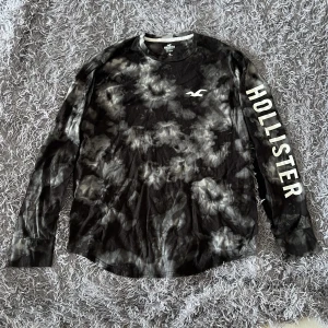Svart tie-dye långärmad tröja från Hollister - Cool långärmad tröja från Hollister i svart och grå tie-dye med stor logga på ärmen och bröstet/ryggen. Tillverkad i 100% bomull, perfekt för dig som gillar streetstyle och vill sticka ut lite extra. Rund halsringning och relaxed fit.