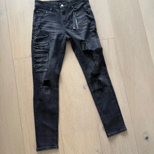 Svarta skinny jeans från Amiri  - Svarta skinny jeans från Amiri med slitningar och trasiga detaljer på benen. Stort AMIRI-tryck i tyg på vänster lår. Jeansen är tillverkade i bomull med inslag av elastan för stretch och har klassisk femficksdesign. Rå och edgy vibe.