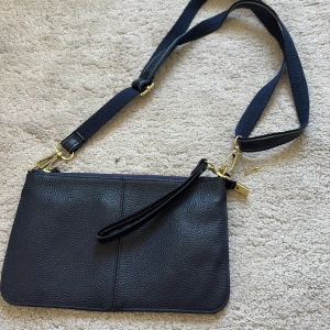 Mörkblå axelväska i skinn med guldiga detaljer - Snygg svart axelväska i äkta skinn med justerbar axelrem och handledsrem. Väskan har guldiga metalldetaljer, dragkedja på baksidan och ett stilrent, minimalistiskt utseende. Perfekt för att bära det viktigaste och passar till många olika outfits.