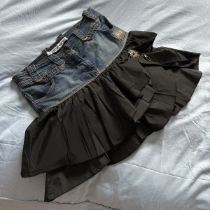 Jeanskjol med svart volang - Unik kortkjol med jeansliv och svart volangkjol. Kjolen har klassiska jeansdetaljer upptill och en cool, asymmetrisk volang i svart tyg nedtill. Fin brosh på sidan! Köpt på en dansk second hand och superfint skick.