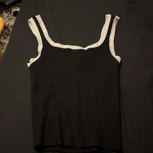 Svart ribbad linnetopp från H&M Divided - Snygg svart ribbad linnetopp från H&M Divided med vita kontrastkanter runt axelband och halsringning. Toppen är ärmlös och har en croppad passform som ger en trendig look. Perfekt att matcha med jeans eller kjol för en cool stil.