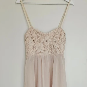 Nudefärgad midiklänning  - Romantisk klänning i färgen nude/beige från Forever21. Överdelen är korsettliknande med blommig spets och kjolen är i tunt svepande meshtyg med asymmetrisk fåll. Tunna axelband. Stängs med dragkedja bak. Passform: Figurnära upptill, faller löst nedtill Skick: Oanvänd med prislapp kvar. Storlek: S