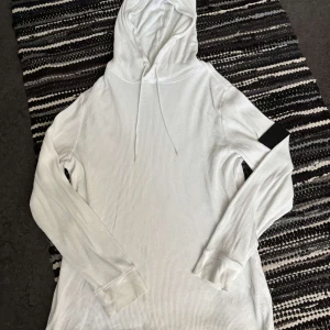Vit hoodie från Calvin Klein Jeans - Vit ribbad hoodie från Calvin Klein Jeans med svart logopatch på ena ärmen. Tröjan har huva med snörning och är tillverkad i mjuk bomull. Perfekt för en chill och stilren look.