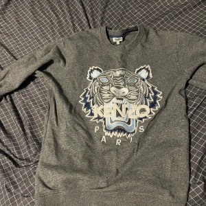 Grå Kenzo sweatshirt med tigertryck - Säljer en grå sweatshirt från Kenzo med ikoniskt broderat tigerhuvud och Kenzo Paris-text i blått och vitt framtill. Tröjan har rund hals, långa ärmar och normal passform. Perfekt statement-plagg för dig som gillar streetstyle.