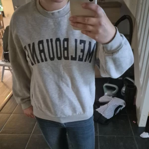 Grå sweatshirt med Melbourne-tryck - Säljer en grå sweatshirt med svart text 'MELBOURNE' över bröstet. Tröjan har rund hals, ribbade muddar och en avslappnad passform. Perfekt för dig som gillar enkel och sportig stil.
