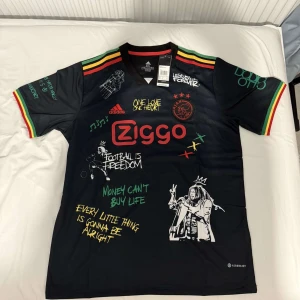 Ajax x Bob Marley Adidas fotbollströja - Unik svart Ajax fotbollströja från Adidas med Bob Marley-tema. Färgglada ränder i rött, gult och grönt på axlar och ärmslut, tryckta citat och illustrationer framtill och baktill. Ziggo-logga på bröstet och Ajax-emblem i rött. Tillverkad i lätt funktionsmaterial.